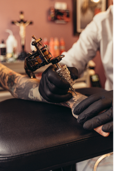 La imagen muestra el proceso de realización de un tatuaje en un entorno profesional. Se puede ver a un tatuador (con guantes negros) utilizando una máquina de tatuar sobre el brazo de una persona que ya tiene tatuajes previos.